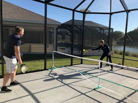 Two people are playing pickleball on a concrete court enclosed by a screened structure. They are dressed in casual athletic attire. One is about to hit a ball with a paddle, while the other watches attentively. The court is outdoors, with grass and a body of water visible in the background under a clear blue sky.