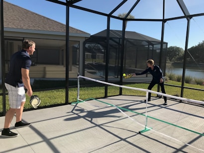 Two people are playing pickleball on a concrete court enclosed by a screened structure. They are dressed in casual athletic attire. One is about to hit a ball with a paddle, while the other watches attentively. The court is outdoors, with grass and a body of water visible in the background under a clear blue sky.