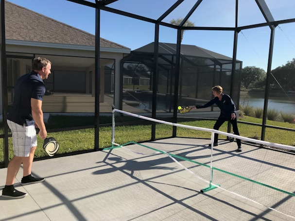Two people are playing pickleball on a concrete court enclosed by a screened structure. They are dressed in casual athletic attire. One is about to hit a ball with a paddle, while the other watches attentively. The court is outdoors, with grass and a body of water visible in the background under a clear blue sky.