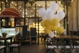A cozy, dimly lit cafe setting with string lights hanging from the ceiling. A cluster of white and yellow balloons is floating near a table. A shop counter with a red awning and a chalkboard menu are visible in the background. There are potted plants and wooden furniture, contributing to a warm ambiance.