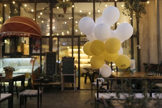 A cozy, dimly lit cafe setting with string lights hanging from the ceiling. A cluster of white and yellow balloons is floating near a table. A shop counter with a red awning and a chalkboard menu are visible in the background. There are potted plants and wooden furniture, contributing to a warm ambiance.