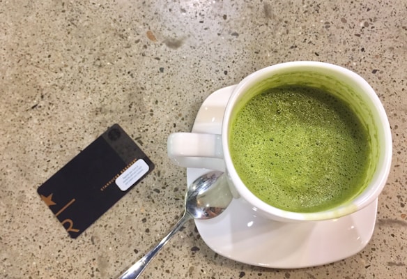 A white ceramic cup filled with frothy green matcha placed on a square white saucer is on a speckled concrete table. Next to the cup, there is a stainless steel spoon and a black card with gold lettering.