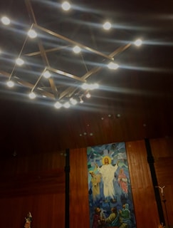 A detailed painting on a wooden wall depicts a religious scene with a central figure dressed in white, surrounded by other figures, possibly disciples, with a radiant halo above the central figure's head. Above the painting, a geometric chandelier with illuminated bulbs hangs from the ceiling.