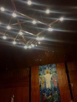 A detailed painting on a wooden wall depicts a religious scene with a central figure dressed in white, surrounded by other figures, possibly disciples, with a radiant halo above the central figure's head. Above the painting, a geometric chandelier with illuminated bulbs hangs from the ceiling.
