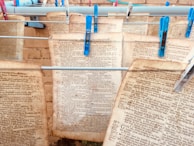 Several pages of old text with faded writing are clipped to clotheslines using blue and grey clothespins. The setup appears to be against a brick or textured wall, hinting at an informal preservation or drying process.