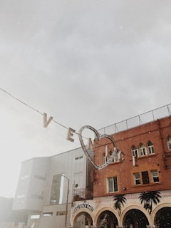 A vintage-style street scene featuring a sign with the word 'Venice' underlined by a heart hanging against a cloudy sky. Below is a building with arched windows and palm tree decorations, identified as the Venice Beach Mart.