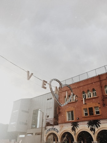 A vintage-style street scene featuring a sign with the word 'Venice' underlined by a heart hanging against a cloudy sky. Below is a building with arched windows and palm tree decorations, identified as the Venice Beach Mart.