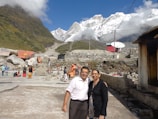 A picturesque mountainous landscape with snow-capped peaks under a clear blue sky. In the foreground, a couple smiles at the camera, while other people are visible in traditional attire among stone structures and satellite dishes. Lush green hills and rocky terrain complete the scene.