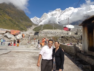 A picturesque mountainous landscape with snow-capped peaks under a clear blue sky. In the foreground, a couple smiles at the camera, while other people are visible in traditional attire among stone structures and satellite dishes. Lush green hills and rocky terrain complete the scene.