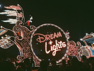 A dazzling parade float adorned with intricate lights forming the words 'Dream Lights.' A character resembling a famous mouse is visible atop the float, surrounded by elaborate light patterns with swirling designs. The crowd is seen watching the parade in the dark, creating a festive atmosphere.