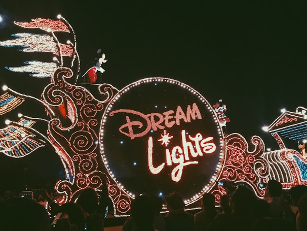A dazzling parade float adorned with intricate lights forming the words 'Dream Lights.' A character resembling a famous mouse is visible atop the float, surrounded by elaborate light patterns with swirling designs. The crowd is seen watching the parade in the dark, creating a festive atmosphere.