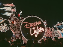 A dazzling parade float adorned with intricate lights forming the words 'Dream Lights.' A character resembling a famous mouse is visible atop the float, surrounded by elaborate light patterns with swirling designs. The crowd is seen watching the parade in the dark, creating a festive atmosphere.
