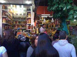 A busy market stall is filled with jars of pickled goods, olives, and spices. People gather around, interacting with the vendors behind the counter. The shelves are stacked with a variety of preserved foods in large glass jars, while the ceiling is adorned with bags of herbs. The environment is lively and bustling as customers browse and purchase items.