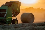 A large green agricultural machine is working in a field during sunset, producing round bales of hay. The atmosphere is filled with a golden haze, and several hay bales are visible in the distance. The machine has wheels and is equipped for processing and bundling the hay.