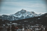 A majestic snow-capped mountain range towers over a small town. The foreground consists of various buildings and rooftops, while the background features rugged peaks under a cloudy sky.