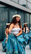 Several women are performing a traditional dance, wearing floral headbands and blue skirts with tropical patterns. They appear to be outdoors, in front of a tall glass building, creating a vibrant and cultural atmosphere.