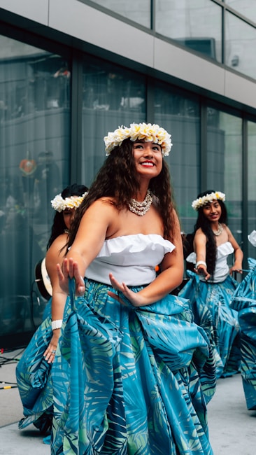 Several women are performing a traditional dance, wearing floral headbands and blue skirts with tropical patterns. They appear to be outdoors, in front of a tall glass building, creating a vibrant and cultural atmosphere.