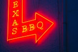 A vibrant red and yellow neon sign in the shape of an arrow pointing to the right, featuring the words 'TEXAS BBQ' with a textured dark blue wall background.