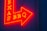 A vibrant red and yellow neon sign in the shape of an arrow pointing to the right, featuring the words 'TEXAS BBQ' with a textured dark blue wall background.