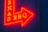 A vibrant red and yellow neon sign in the shape of an arrow pointing to the right, featuring the words 'TEXAS BBQ' with a textured dark blue wall background.