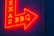 A vibrant red and yellow neon sign in the shape of an arrow pointing to the right, featuring the words 'TEXAS BBQ' with a textured dark blue wall background.