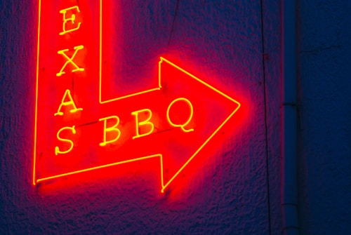 A vibrant red and yellow neon sign in the shape of an arrow pointing to the right, featuring the words 'TEXAS BBQ' with a textured dark blue wall background.