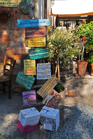 Colorful handwritten signs display a variety of food and drink offerings at an outdoor cafe, surrounded by green plants and a rustic setting. Two cardboard boxes are placed on the ground nearby.
