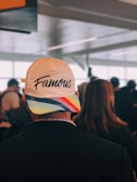A person wearing a white baseball cap with the word 'Famous' written on it, featuring a colorful rainbow pattern on the brim. The setting appears to be an indoor location with several other people in the background, likely a public or transportation space.