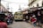 A busy street scene with a colorful bus driving through a narrow road lined with small shops and stalls on both sides. People are walking on the street, some browsing items for sale. The bus is decorated with vibrant yellow and green colors and has a destination sign at the front. Overhead wires crisscross above, and there are various signs and goods displayed outside the shops.