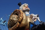 A large, colorful carnival float with a caricature of a distinguished older man wearing a top hat. He is depicted with a pot overflowing with gold coins. The backdrop includes blue and teal decorative elements, suggesting a festive or parade setting.