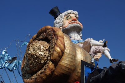 A large, colorful carnival float with a caricature of a distinguished older man wearing a top hat. He is depicted with a pot overflowing with gold coins. The backdrop includes blue and teal decorative elements, suggesting a festive or parade setting.