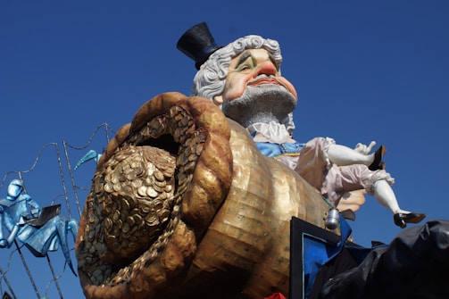 A large, colorful carnival float with a caricature of a distinguished older man wearing a top hat. He is depicted with a pot overflowing with gold coins. The backdrop includes blue and teal decorative elements, suggesting a festive or parade setting.