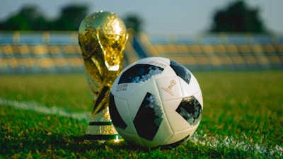 A gold trophy and a black-and-white soccer ball are placed on a green grass field. The background shows blurred stadium seating, suggesting a sports venue.