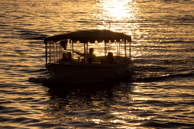 A small boat with a canopy glides across a shimmering water surface during sunset. Silhouettes of people are visible aboard, creating a peaceful and picturesque scene.