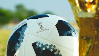 A soccer ball designed for the FIFA World Cup 2018 with a pixelated pattern is positioned next to a golden trophy. The background is blurred, suggesting an outdoor setting with green and brown hues.