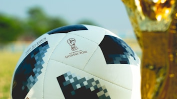 A soccer ball designed for the FIFA World Cup 2018 with a pixelated pattern is positioned next to a golden trophy. The background is blurred, suggesting an outdoor setting with green and brown hues.
