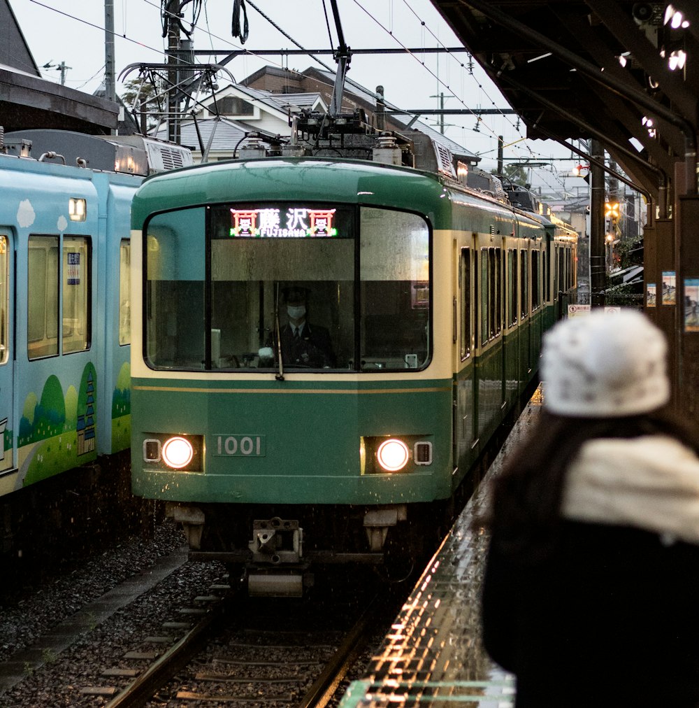 woman standing on waiting shed facing speeding green and white train