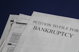 Documents are laid out on a surface, with the largest sheet displaying the text 'Petition to File for Bankruptcy'. Several forms and paperwork are visible underneath, suggesting a formal or legal context.