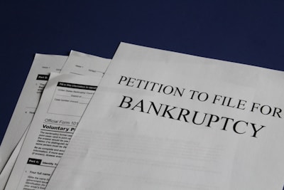 Documents are laid out on a surface, with the largest sheet displaying the text 'Petition to File for Bankruptcy'. Several forms and paperwork are visible underneath, suggesting a formal or legal context.