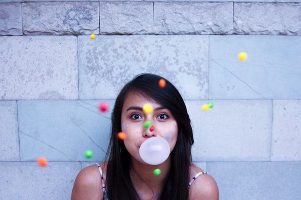 a woman with light skin and black hair standing against a wall, facing the viewer, and blowing a gum bubble