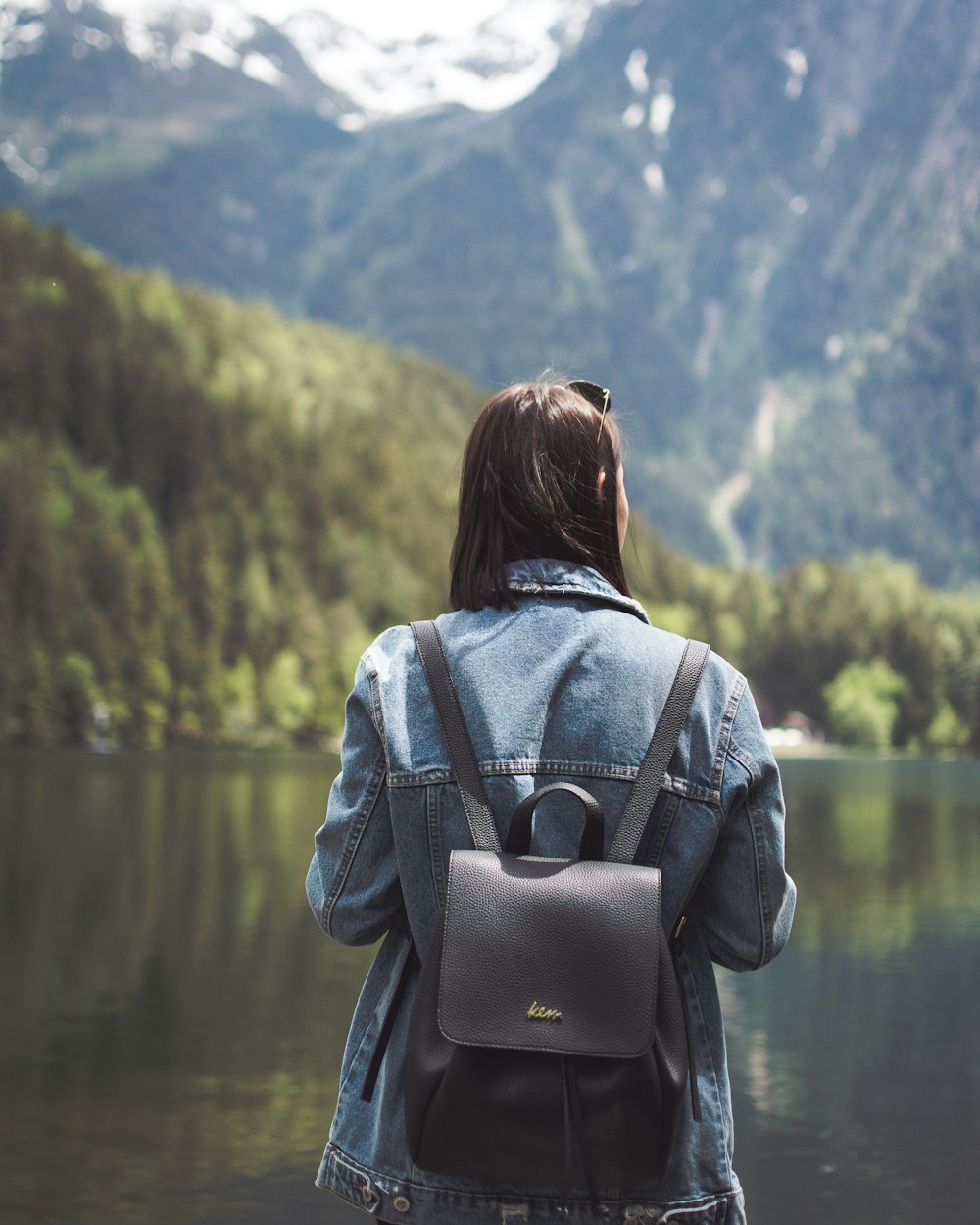 selective focus photography of woman wearing blue denim jacket and black bucket backpack near lagoon at daytime