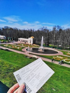A scenic park area features a large circular fountain surrounded by well-maintained lawns and ornate flowerbeds. In the foreground, a person is holding two entrance tickets. The sky is bright blue, indicating a clear and sunny day.