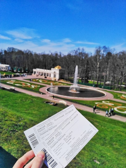 A scenic park area features a large circular fountain surrounded by well-maintained lawns and ornate flowerbeds. In the foreground, a person is holding two entrance tickets. The sky is bright blue, indicating a clear and sunny day.