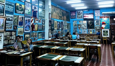 The interior of a cozy restaurant is decorated with numerous framed photographs and posters on the walls, creating a nostalgic atmosphere. Wooden tables and chairs are arranged neatly throughout the room, with patrons sitting and enjoying their meals. The lighting is soft, complementing the vibrant blue walls.