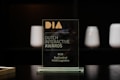 A glass award plaque etched with the text 'Dutch Interactive Awards' and other smaller inscriptions. It is placed on a black reflective surface with a blurred dark background and some softly lit cylindrical objects visible in the backdrop.