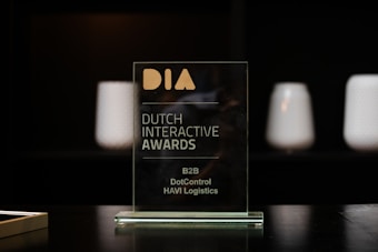 A glass award plaque etched with the text 'Dutch Interactive Awards' and other smaller inscriptions. It is placed on a black reflective surface with a blurred dark background and some softly lit cylindrical objects visible in the backdrop.