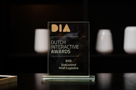 A glass award plaque etched with the text 'Dutch Interactive Awards' and other smaller inscriptions. It is placed on a black reflective surface with a blurred dark background and some softly lit cylindrical objects visible in the backdrop.