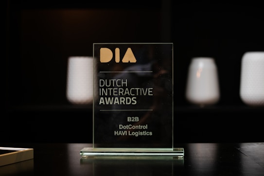 A glass award plaque etched with the text 'Dutch Interactive Awards' and other smaller inscriptions. It is placed on a black reflective surface with a blurred dark background and some softly lit cylindrical objects visible in the backdrop.