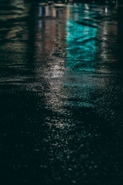 Wet pavement reflects colorful lights, with prominent shades of turquoise and subtle hints of other hues. The textured surface adds a sense of depth, creating a serene yet mysterious ambiance.