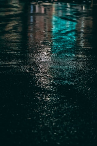 Wet pavement reflects colorful lights, with prominent shades of turquoise and subtle hints of other hues. The textured surface adds a sense of depth, creating a serene yet mysterious ambiance.